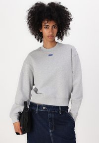 Gray sweatshirt with a crew neckline, featuring a blue logo on the chest, paired with a denim skirt and black pouch at the waist.