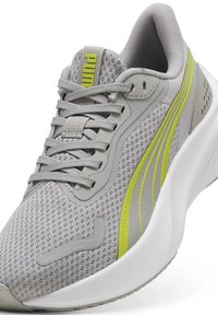 Zapatilla deportiva gris con parte superior de malla, acentos grises y amarillos, diseño texturizado, cierre de cordones y suela blanca gruesa. Presenta un logo en la lengüeta.