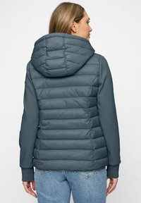Frau trägt eine blau-graue gesteppte Daunenjacke mit Kapuze und einfarbigen blauen Ärmeln, kombiniert mit hellblauen Jeans, mit dem Rücken zur Kamera.