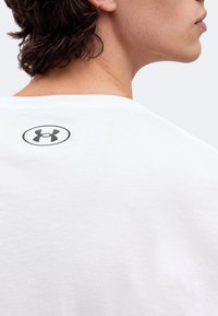 Gros plan sur le cou et l'épaule d'une personne portant une chemise blanche avec un logo noir Under Armour dans le haut du dos.