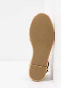 Semelle d'une espadrille beige à semelle en caoutchouc avec une adhérence texturée et un bord en jute tissé, placée debout contre un coin blanc.