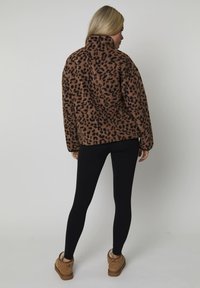 Donna vestita con una giacca in pile leopardata marrone, leggings neri e scarpe foderate in shearling color sabbia, in piedi di fronte a un muro grigio chiaro.