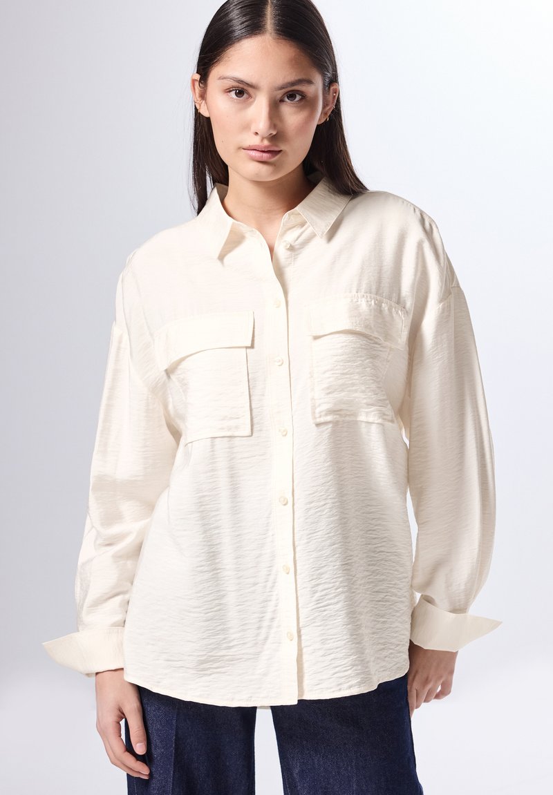 Camisa de botones color crema con dos bolsillos en el pecho, ajuste relajado, tela texturizada y mangas largas con puños doblados.
