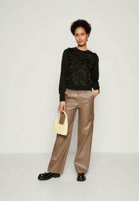 Žena nosi crni strukturni pulover, smeđe široke hlače, crne robusne cipele, drži malu bež torbicu i stoji nasuprot jednoličnoj pozadini.