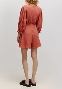 Terracotta gekleurde shorts set met lange, gepofte mouwen en een gestrikte taille, met een relaxte pasvorm en een eenvoudige halslijn, gedragen met zwarte sandalen.