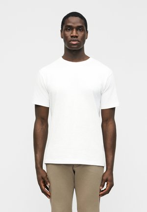 Jeune homme noir debout, face à l'objectif, portant un T-shirt blanc uni à manches courtes et un pantalon beige, sur un fond uni.