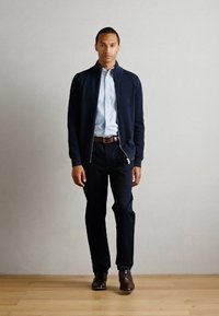 Cárdigan azul marino con cremallera sobre una camisa de botones azul claro, combinado con pantalones oscuros y zapatos de cuero marrón, sobre un suelo de madera clara.