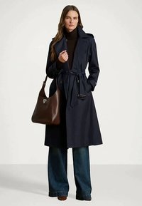 Γυναίκα με ναυτικό μπεζ παλτό με ζώνη, σκούρο ζιβάγκο, τζιν φαρδιάς γραμμής, καφέ παπούτσια, και κρατώντας μια μεγάλη καφέ δερμάτινη τσάντα.