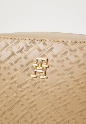 Surface en cuir beige avec un motif géométrique embossé et un petit logo métallique doré comprenant les lettres T et H entrelacées.