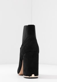 Stivale alla caviglia in suede nero con tacco a blocco alto, caratterizzato da pannelli laterali in elastico e un accento metallico vicino alla base del tacco.