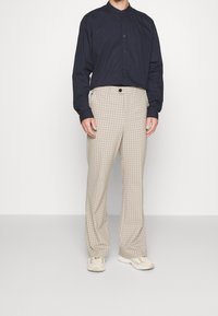 Muž v tmavě modré košili na knoflíky, béžových kostkovaných kalhotách a béžových teniskách stojí před jednoduchým bílým pozadím.