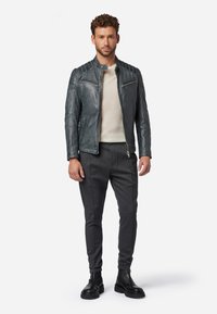 Grijze leren jas met geribbelde schouderdetails, gecombineerd met een beige trui en donkergrijze broek. Zwarte enkellaarzen maken de outfit compleet.