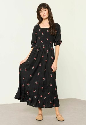 Donna con capelli lunghi e scuri che indossa un lungo vestito nero con stampe floreali rosa e scarpe basse beige, in piedi su un pavimento grigio contro un muro bianco.