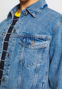 Světlá modrá džínová bunda s límcem, dvěma náprsními kapsami a druky na zapínání. Má viditelné švy a texturovanou látku.