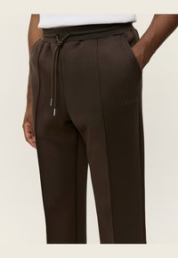 Braune Sporthose mit glatter Textur, elastischem Taillenbund mit Kordelzug und Seitentaschen. Mit dem bedruckten Text "LES DRA.".