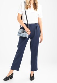 Donna che indossa una maglietta bianca a maniche corte, pantaloni blu navy a vita alta, scarpe con tacco nere, mentre tiene una piccola borsa a spalla grigia trapuntata con tracolla a catena.