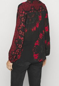 Osoba ubrana w czarną i czerwoną bluzę z długim rękawem o wzorach kwiatowych i tie-dye, w zestawieniu z czarnymi dżinsami, odwrócona plecami do kamery.