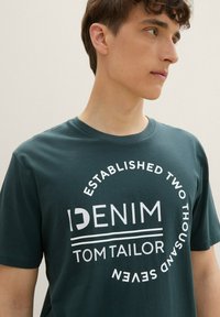 Camiseta de algodón verde con cuello redondo, que presenta texto blanco en contraste: "DENIM" y "TOM TAILOR", con diseño circular y año de establecimiento.