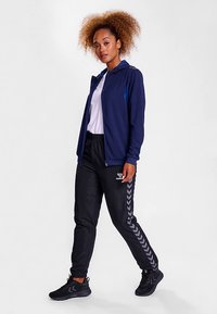 Marineblauer Kapuzenpullover mit Reißverschluss über einem weißen Hemd, kombiniert mit schwarzen Jogginghosen mit grauem Chevron-Muster an den Seiten. Schwarze Turnschuhe vervollständigen das Outfit.