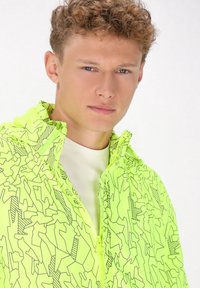 Lys neon gul jakke med hætte, der har et geometrisk sort linjemønster, lynlås lukning og en let, glat tekstur.
