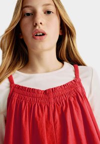 Abito rosso di cotone con tessuto arricciato e spalline regolabili, indossato sopra una maglietta bianca a maniche lunghe a costine, che mostra una texture liscia e un design semplice.