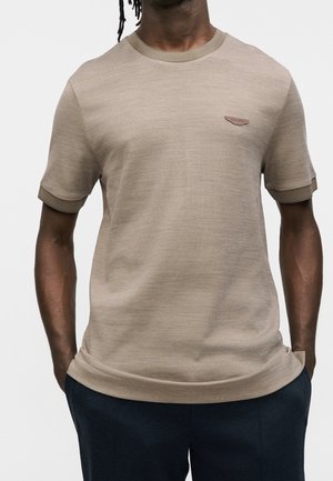 Homme portant une chemise en tricot beige à manches courtes avec col et poignets côtelés, et un petit logo en forme d'aile sur la poitrine, mains dans les poches d’un pantalon bleu marine.