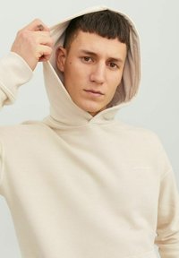Beżowa bluza z kapturem ze sznurkiem, miękka w dotyku, z małym logo na klatce piersiowej. Model pokazuje wewnętrzną wyściółkę.