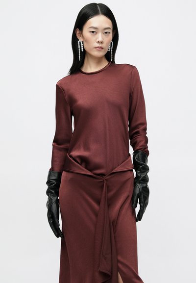 Top de cetim bordeaux com mangas longas, apresentando um detalhe em nó na cintura. Usado com luvas de couro pretas, design elegante e ajustado.