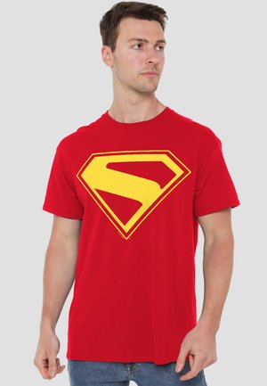 Young man wearing a red t-shirt with a large yellow stylized "S" emblem, looking to the right against a plain background.