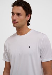 Vit t-shirt med kort ärm i slät tyg, med en liten svart chevronlogotyp på vänster bröst. Rund halsringning, normal passform.