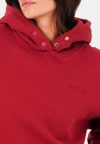 Un sweat à capuche rouge en tissu doux, doté d'une capuche avec cordon de serrage, de boutons-pression et d'un logo de la marque brodé sur la poitrine. Texture lisse.