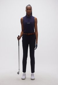 Γυναίκα που στέκεται κρατώντας ένα γκολφ κλαμπ, φοράει μπλε σκούρο αμάνικο πόλο, μαύρο παντελόνι, γάντι γκολφ και λευκά παπούτσια γκολφ.