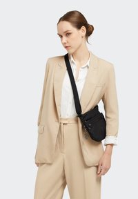 Black crossbody bag with embossed pattern, adjustable strap, and multiple zip pockets, worn over a cream suit and white shirt.