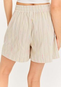 Béžové šortky s vertikálnymi viacfarebnými pruhmi, elastickým pásom, nosené osobou zobrazenou od pásu nadol na bielom pozadí.