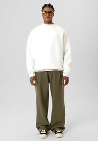 Witte oversized sweatshirt met een hoge hals, olijfgroene broek met wijde pijpen en zwarte sneakers, gecombineerd met geelgetinte zonnebrillen.