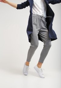 Grå sweatpants med snörning, kombinerat med en vit skjorta och en lång mörkblå kappa. Vita sneakers fulländar outfiten.