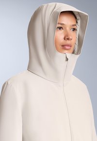 Jakna krem boje s kapuljačom, glatka, mat završna obrada. Ima visok ovratnik i središnji patentni zatvarač, dizajnirana za otpornost na vremenske uvjete.