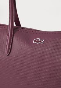 Bordo spalvos rankinė su tekstūruotu paviršiumi, plonomis rankenėlėmis ir mažu baltu siuvinėjimu su lacoste logotipu priekyje.