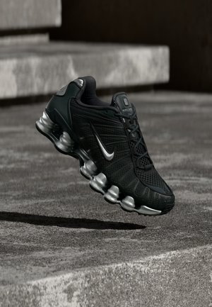 Fekete Nike futócipő ezüst talppal és logóval, beton lépcsők felett lebegve városi környezetben.