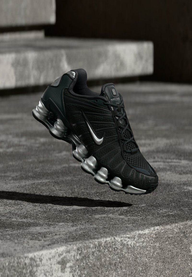 Zapatilla de correr Nike negra con suela y logo plateados, suspendida sobre escalones de concreto en un entorno urbano.