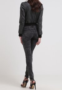 Donna in piedi con la schiena rivolta verso la fotocamera, che indossa una giacca di pelle nera, jeans skinny scuri e sandali neri con tacco alto.