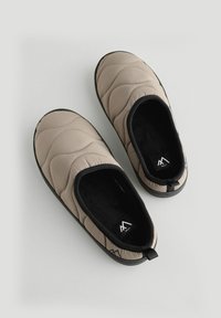 Zapatillas acolchadas beige con acentos negros, suave forro interior, punta redondeada y suela de goma texturizada para agarre. Incluye una pestaña trasera para ayudar a calzarlas.