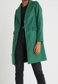 Abrigo verde de mezcla de lana con cuello de solapa, cierre con un solo botón y bolsillos laterales, combinado con jeans negros. Textura suave y ajuste entallado.