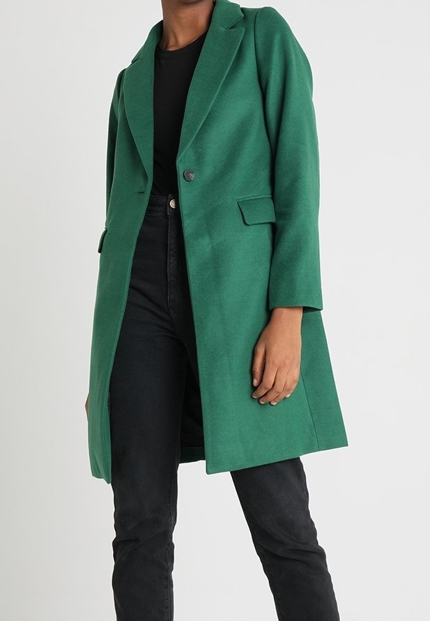 Abrigo verde de mezcla de lana con cuello de solapa, cierre con un solo botón y bolsillos laterales, combinado con jeans negros. Textura suave y ajuste entallado.