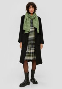 Long manteau noir, écharpe texturée verte, robe à carreaux avec des accents verts et noirs, associée à des bottes noires épaisses et des collants.