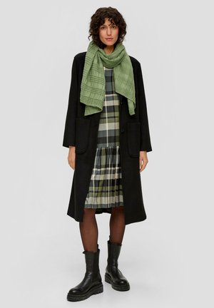 Long manteau noir, écharpe texturée verte, robe à carreaux avec des accents verts et noirs, associée à des bottes noires épaisses et des collants.