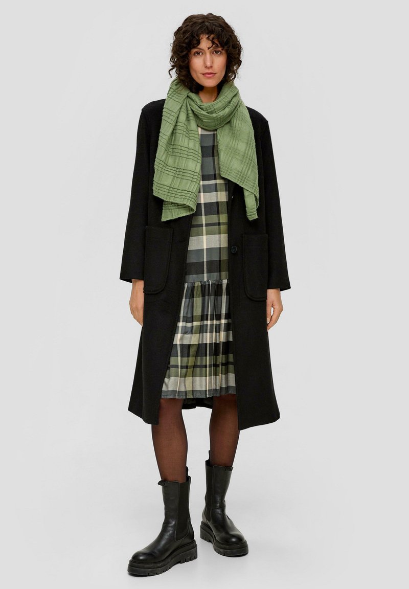 Long manteau noir, écharpe texturée verte, robe à carreaux avec des accents verts et noirs, associée à des bottes noires épaisses et des collants.