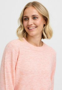 Maglione a maglia rosa chiaro, scollatura rotonda, texture morbida con sottili variazioni. Il modello ha i capelli corti biondi e sorride leggermente.