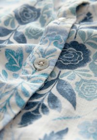 Camicia a maniche corte con bottoni in tessuto leggero, decorata con motivi floreali in blu e turchese. Presenta un bottone rotondo di colore chiaro e una texture morbida.