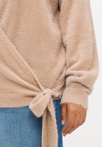 Μπεζ πλεκτό πουλόβερ με απαλή υφή, διακοσμημένο με κόμπο στη μέση και φαρδιές μανσέτες, συνδυασμένο με μπλε τζιν.